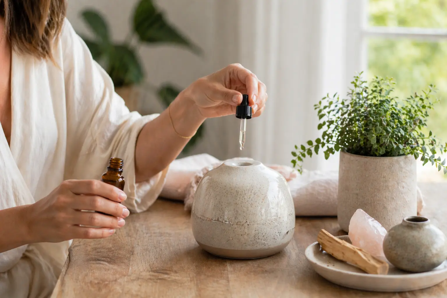 Hands adding essential oil drops into a ceramic diffuser in bright morning light